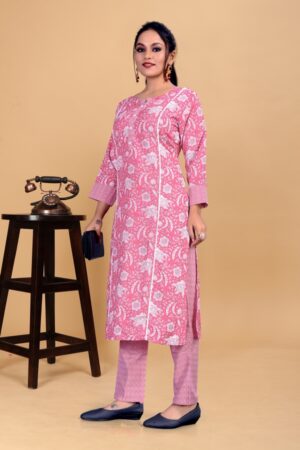 A woman stands in a pink patterned kurta with matching pants, holding a small purse, against a beige background.